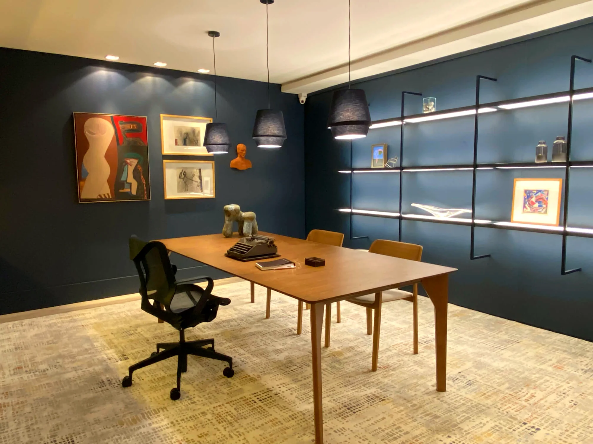 Home office da mostra Refúgio da Alma, com mesa de trabalho em madeira, estante iluminada e obras de arte na parede
